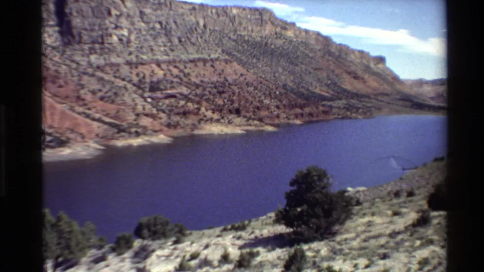 Western USA 1980 home-movie frame showing a blue reservoir below layered canyon walls and sparse desert vegetation.