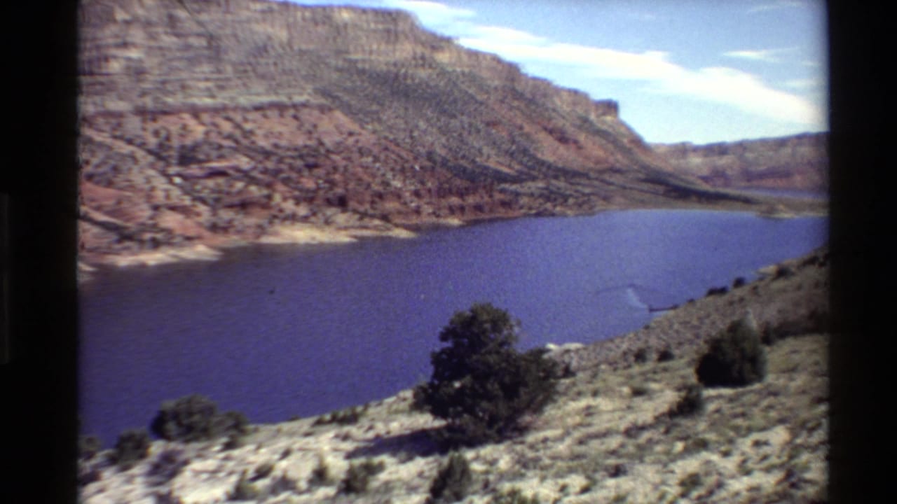 1980 Western USA 8mm frame of a canyon reservoir, foreground tree, rocky slope, and distant red rock walls under daylight.