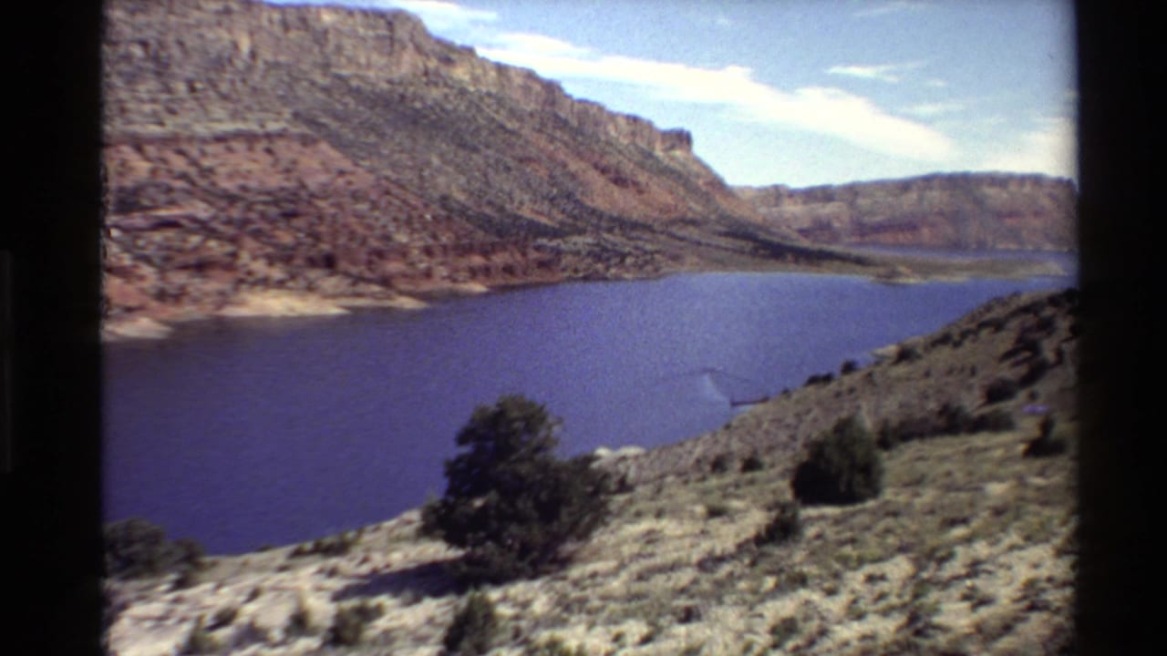 Western USA 1980 home-movie view from a rocky overlook toward a blue canyon reservoir and layered desert cliffs.