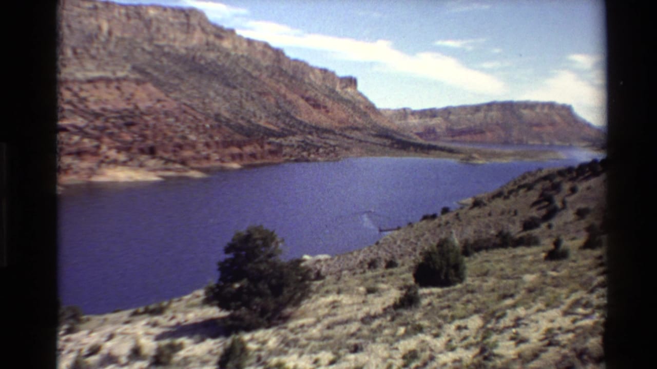 1980 Western U.S. 8mm view of a deep blue river below red canyon cliffs, seen from a scrub-covered overlook.