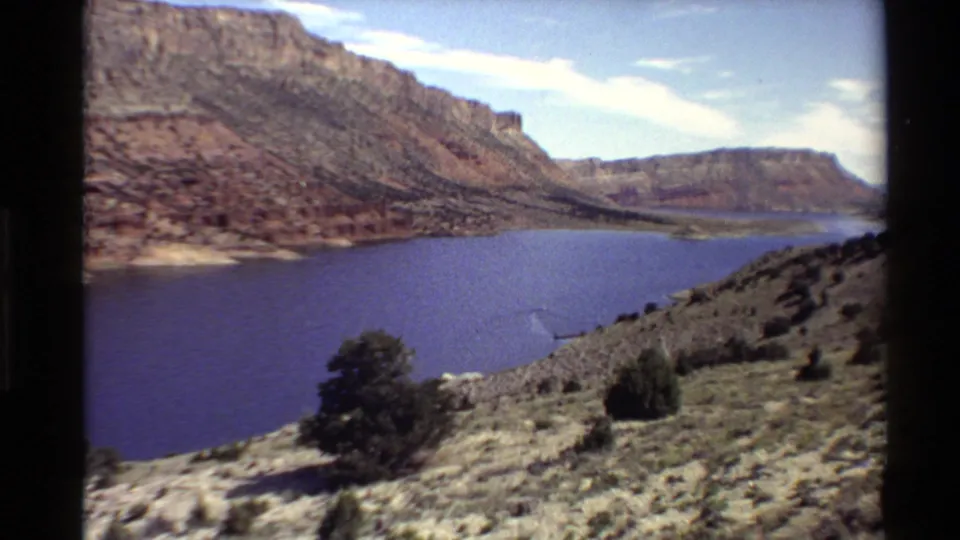 1980 Western U.S. 8mm view of a deep blue river below red canyon cliffs, seen from a scrub-covered overlook.