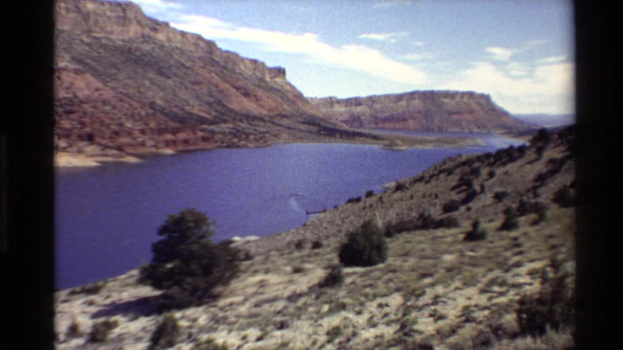 Wide canyon river view with dry hillside plants and cliffs beyond the water.
