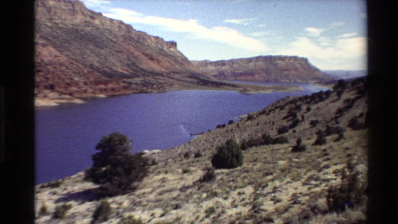 8mm travel footage frame of a canyon gorge with blue water, red rock cliffs, and sparse Western U.S. desert vegetation.