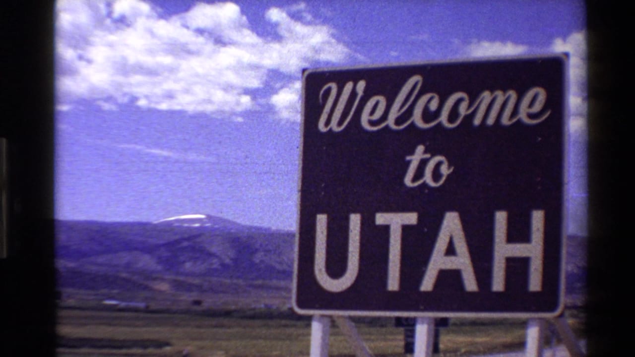 Welcome to Utah sign beside open grassland with mountains and clouds behind it.