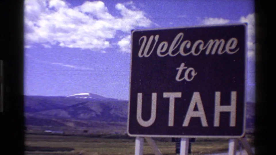 Bright 1980 8mm frame of a Welcome to Utah roadside sign with open grassland, distant mountains, and a blue sky with clouds.