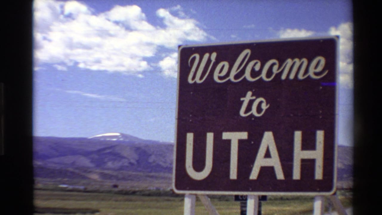 Closer 1980 8mm frame of the Welcome to Utah sign filling much of the view, backed by grassland, mountains, and scattered clouds.