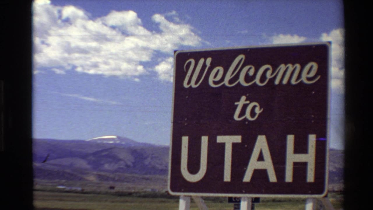 1980s 8mm frame showing a Welcome to Utah highway sign against distant mountains and a partly cloudy blue sky.