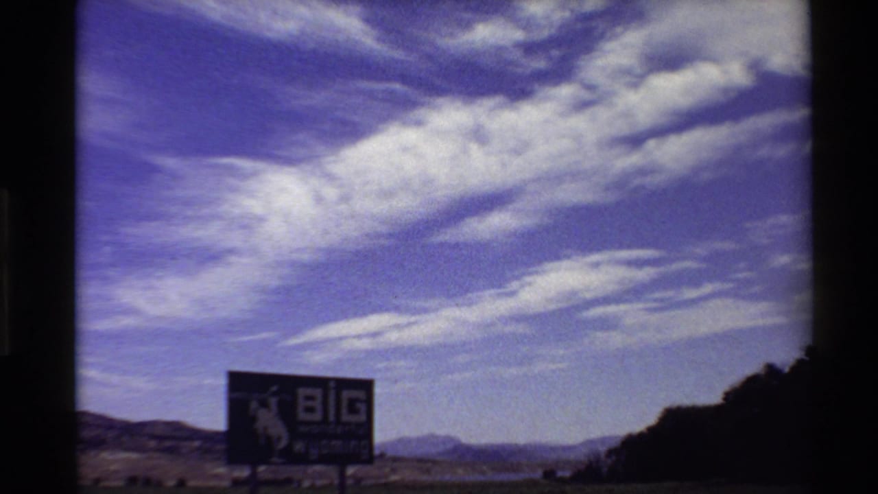 Wide 8mm frame showing the Big Wyoming billboard, open roadside terrain, distant mountains, and a cloud-filled blue-purple sky.