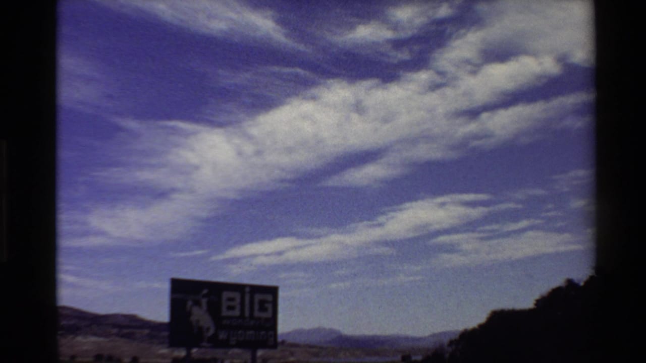 1980s 8mm Western USA view of the "BIG wonderful wyoming" sign, distant mountains, and wide blue sky with white cloud bands.