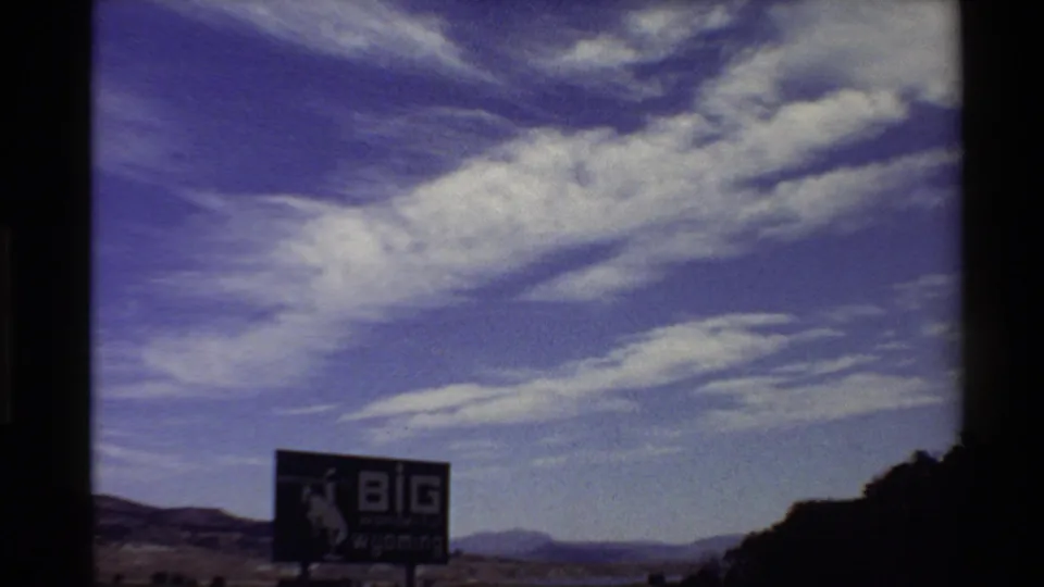 1980s 8mm Western USA view of the "BIG wonderful wyoming" sign, distant mountains, and wide blue sky with white cloud bands.