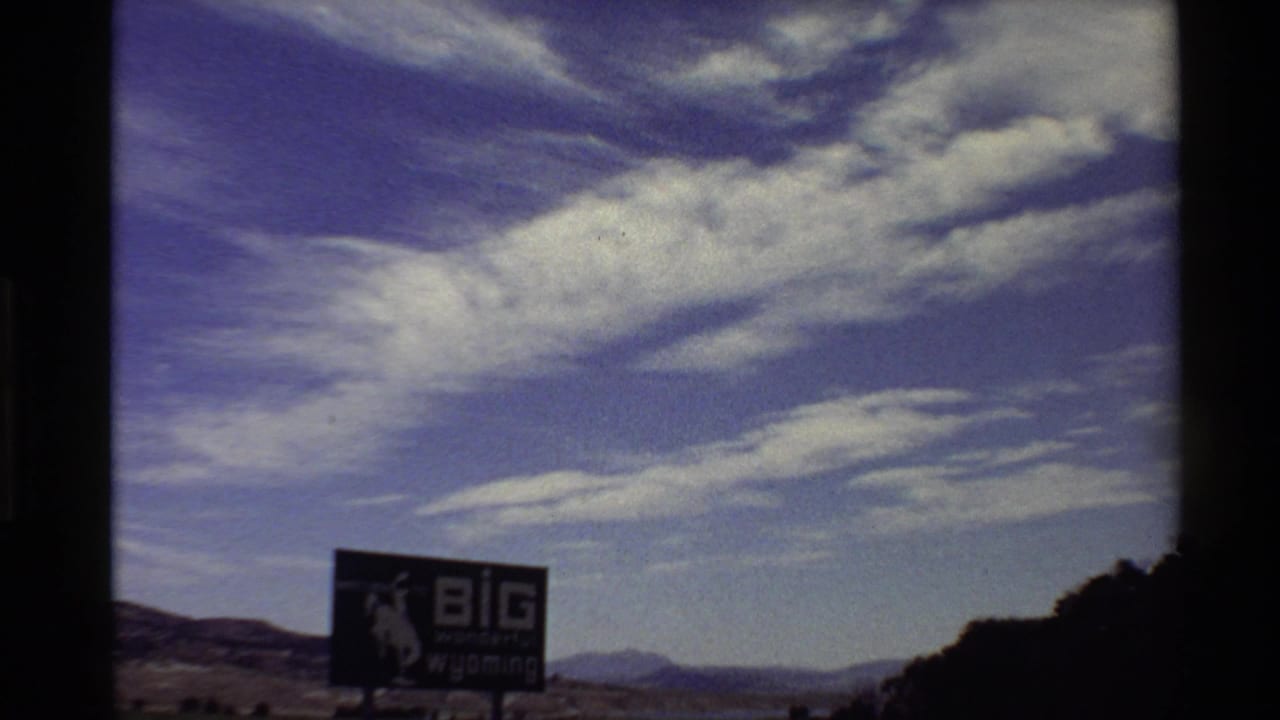 Western USA 8mm frame from 1980 showing a large Wyoming-themed roadside billboard below layered clouds and distant hills.