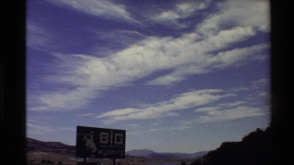 Western USA 8mm frame from 1980 showing a large Wyoming-themed roadside billboard below layered clouds and distant hills.