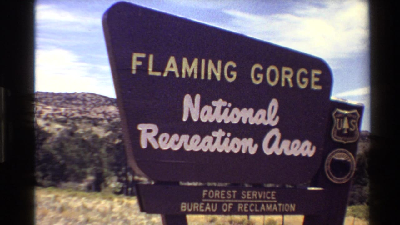 Centered 1980 Western U.S. home-movie frame of the Flaming Gorge National Recreation Area sign, with arid hills and sky in the background.