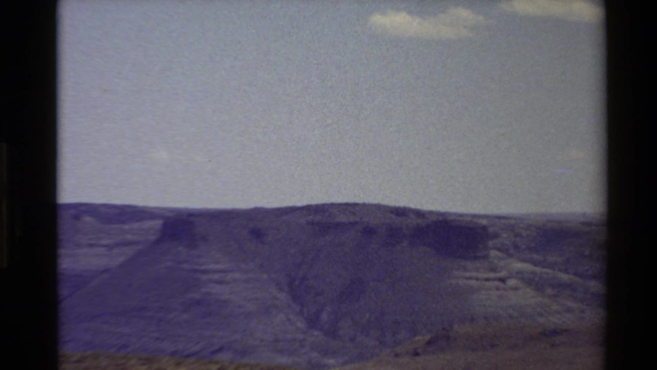 Broad mesa-like ridge and canyon slope under a large pale sky with dark vertical film borders.