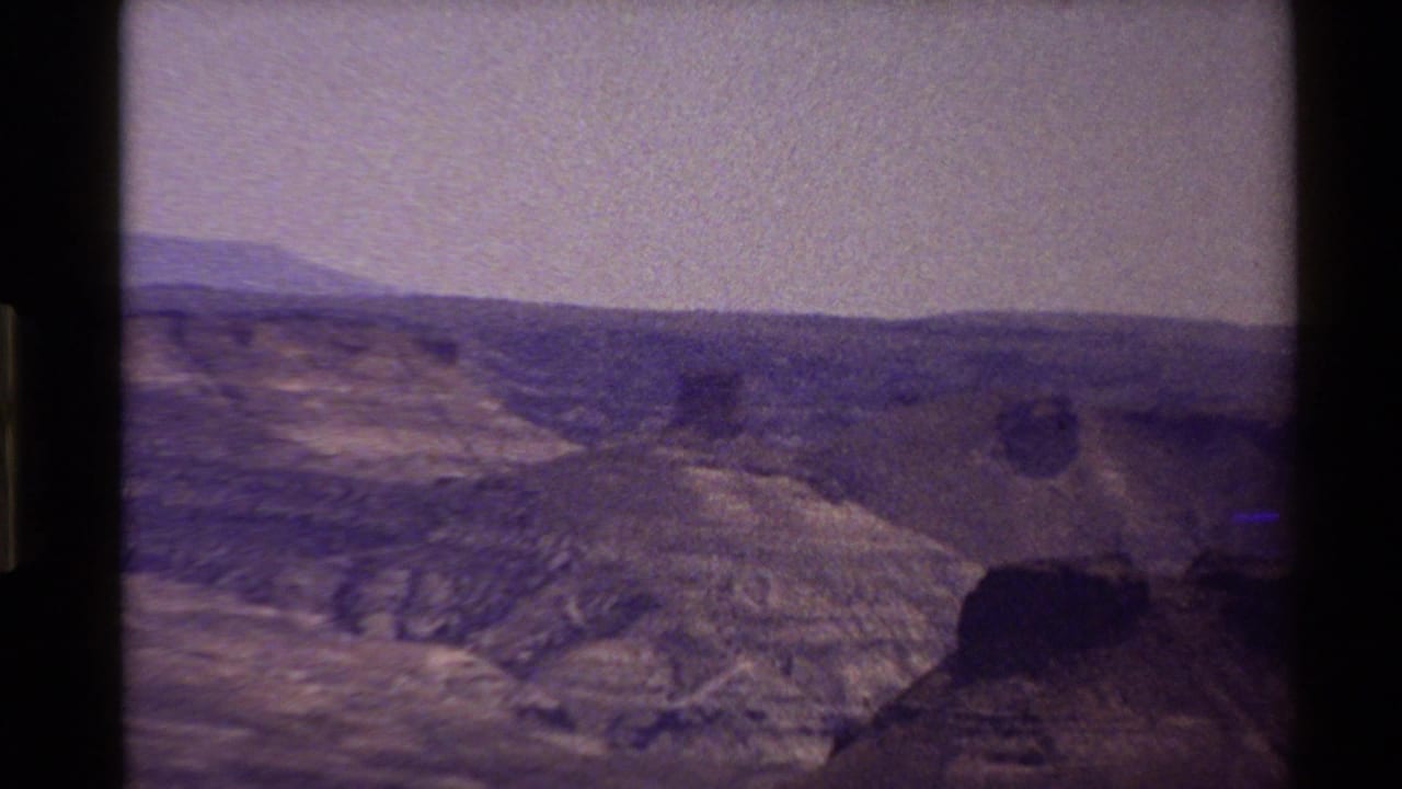 Grainy 8mm view of rocky Western USA mountain terrain under a pale purple sky, framed by dark film edges.