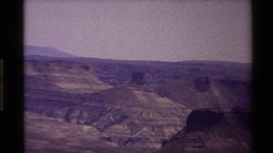 Grainy 8mm view of rocky Western USA mountain terrain under a pale purple sky, framed by dark film edges.