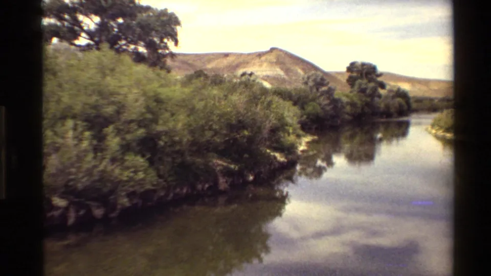 Western USA 8mm home-movie view of a calm river or lake edge bordered by shrubs, trees, and low hills.