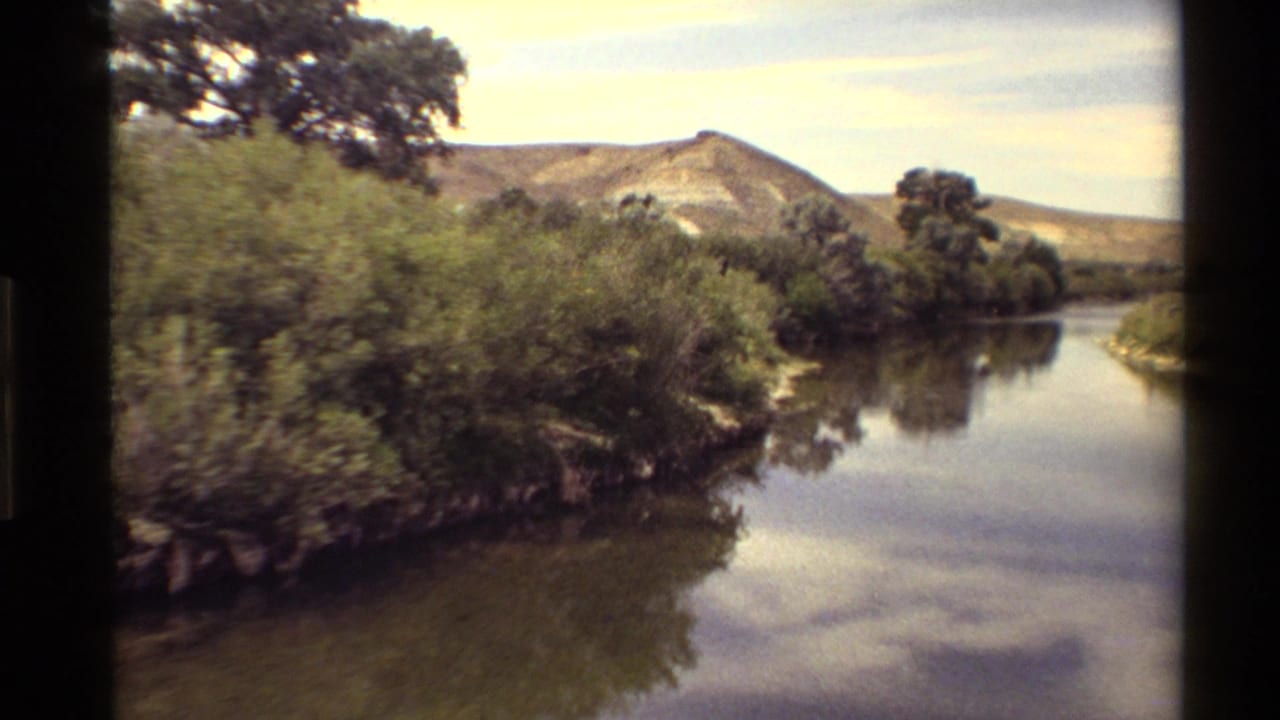 Vintage 8mm Western USA landscape showing brush along a calm waterway, mature trees, and a rounded hill beyond.