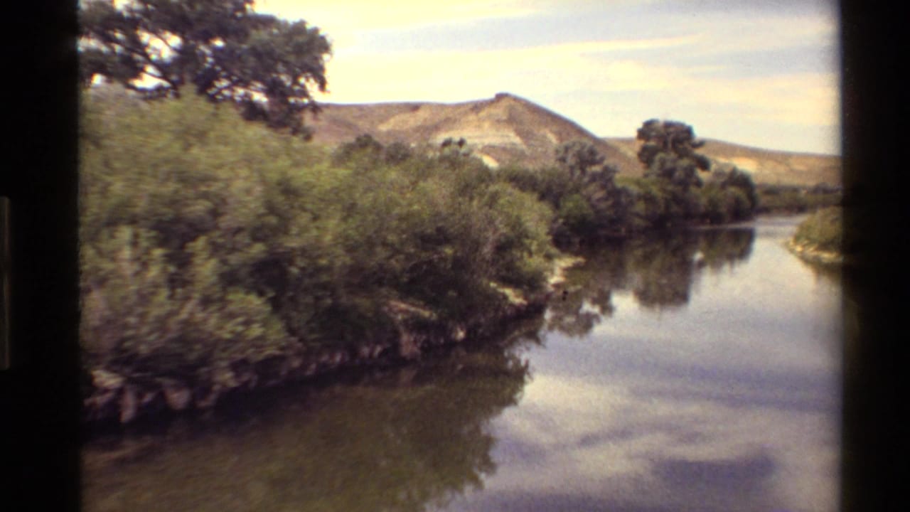 Western USA 8mm frame of a quiet waterside scene with shrubs, reflected clouds, trees, and distant hills.