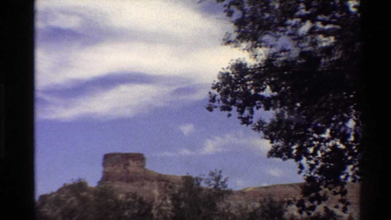 1980 Western U.S. home-movie view of a distant flat-topped rock formation beneath wispy clouds, with tree foliage at the frame edge.
