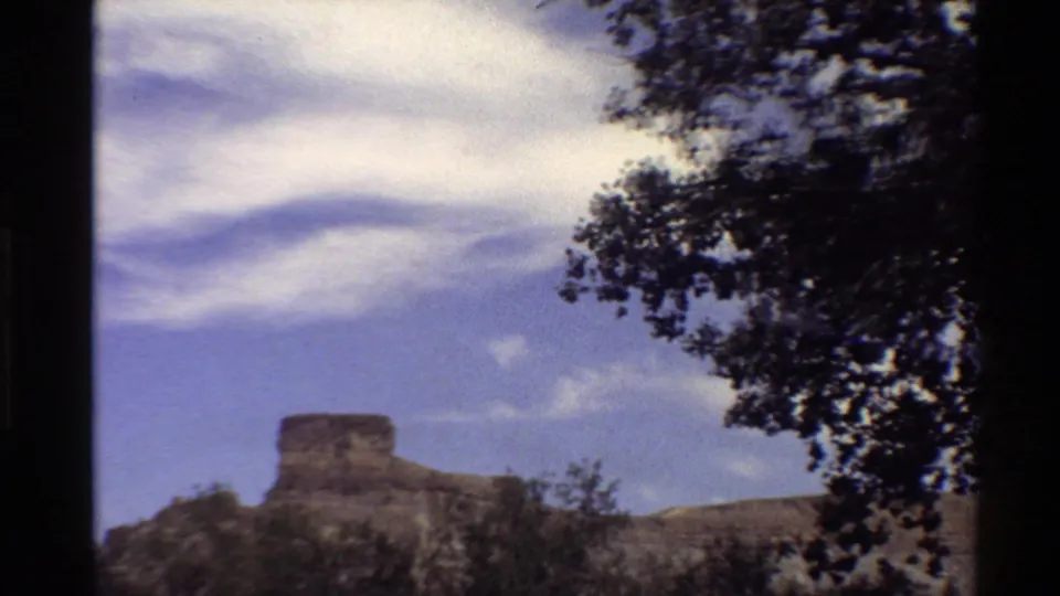 1980 Western U.S. home-movie view of a distant flat-topped rock formation beneath wispy clouds, with tree foliage at the frame edge.