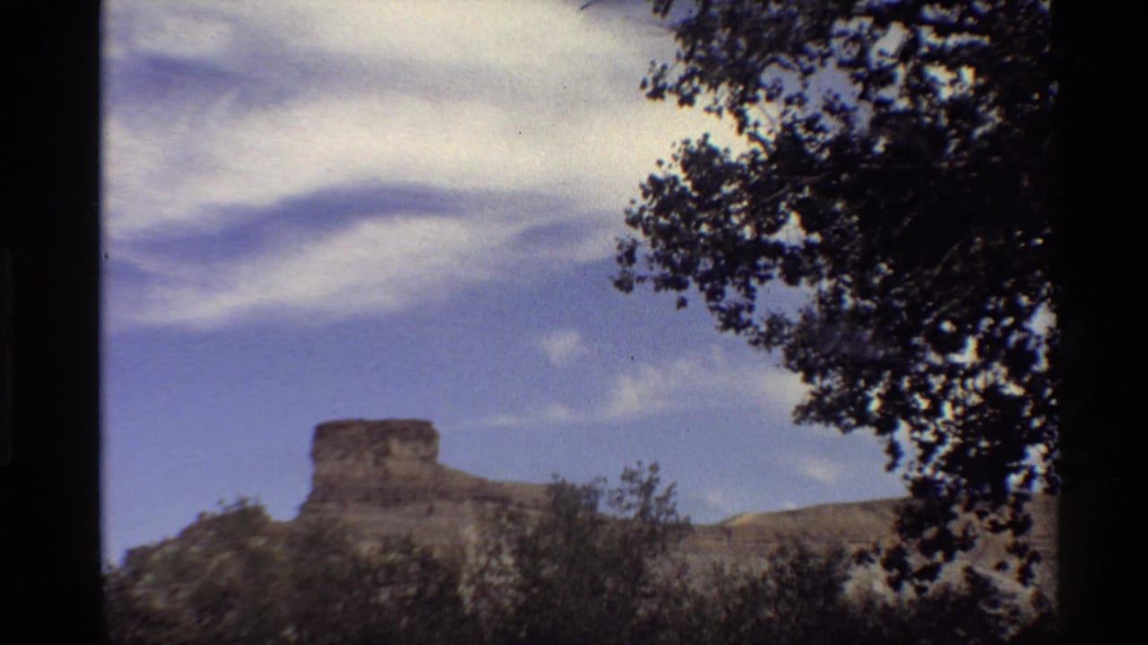 A second handheld 8mm view holds on the Western U.S. rock formation, with brushy terrain below and dark foliage along the upper right.