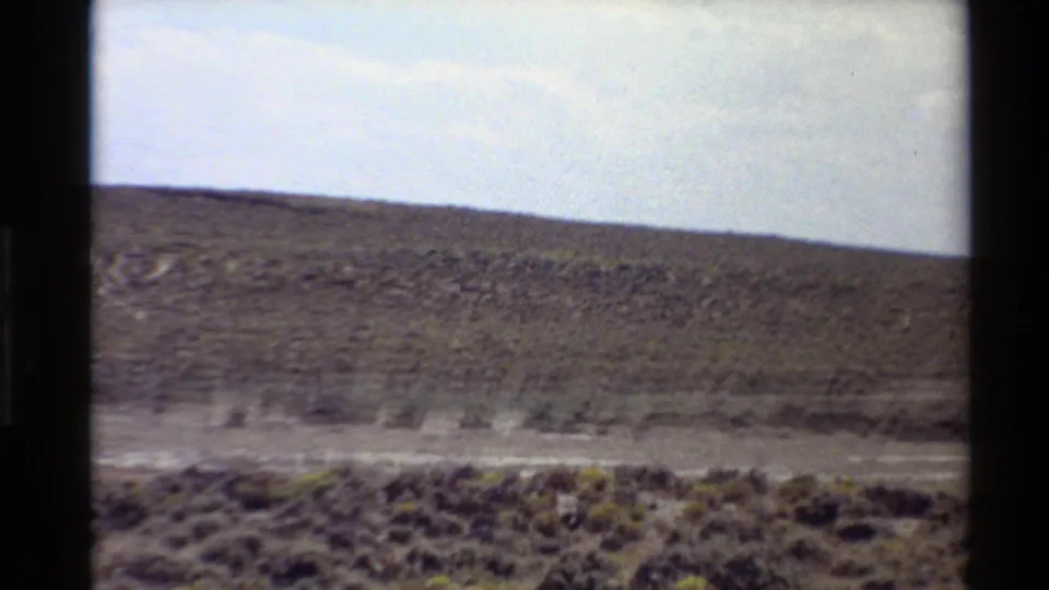 1980 Western U.S. home-movie frame showing small antelope running across open scrub grassland beneath low hills.