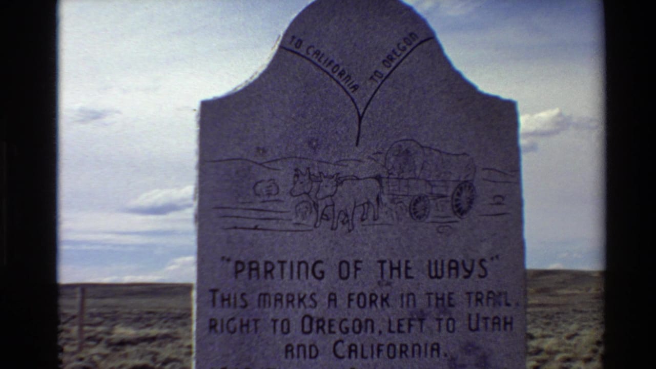 1980 8mm frame of the Parting of the Ways trail marker, showing text, wagon carving, cloudy sky, and Western USA plains.