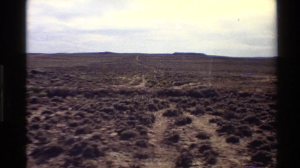 Western U.S. desert view on 8mm film, with low brush, a pale winding track, and flat distant hills under high light.
