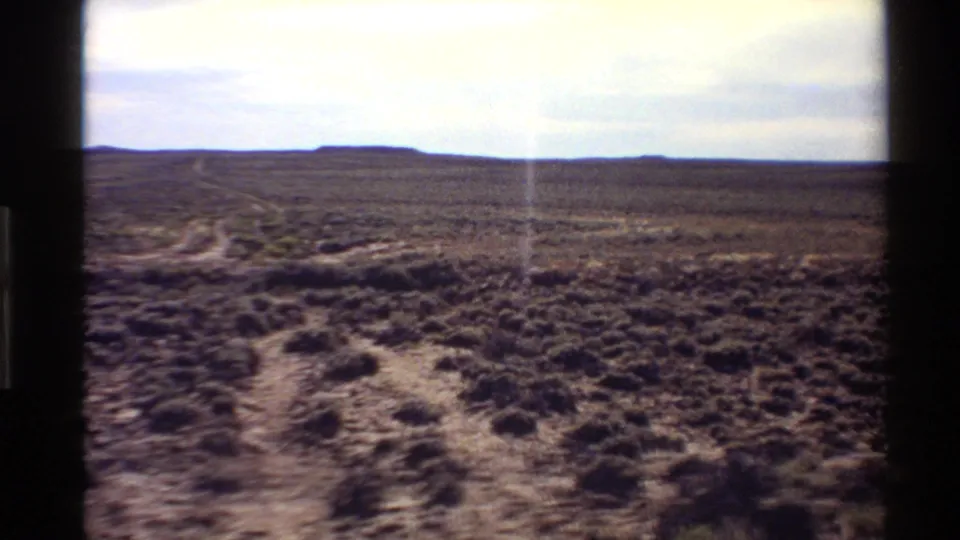 1980 Western U.S. 8mm view of open scrub desert with a faint dirt path crossing the foreground beneath a pale sky.
