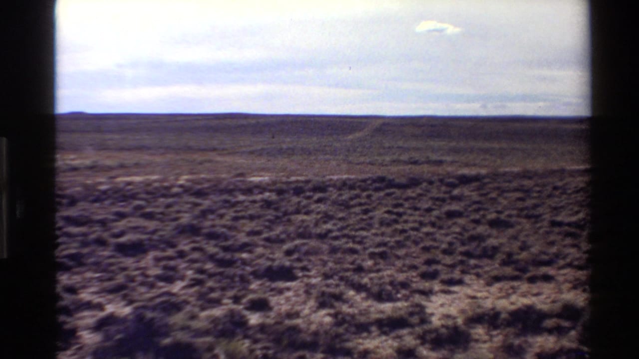 Western U.S. 1980 home-movie view of scrub-covered desert with a faint track and distant horizon under a pale clouded sky.