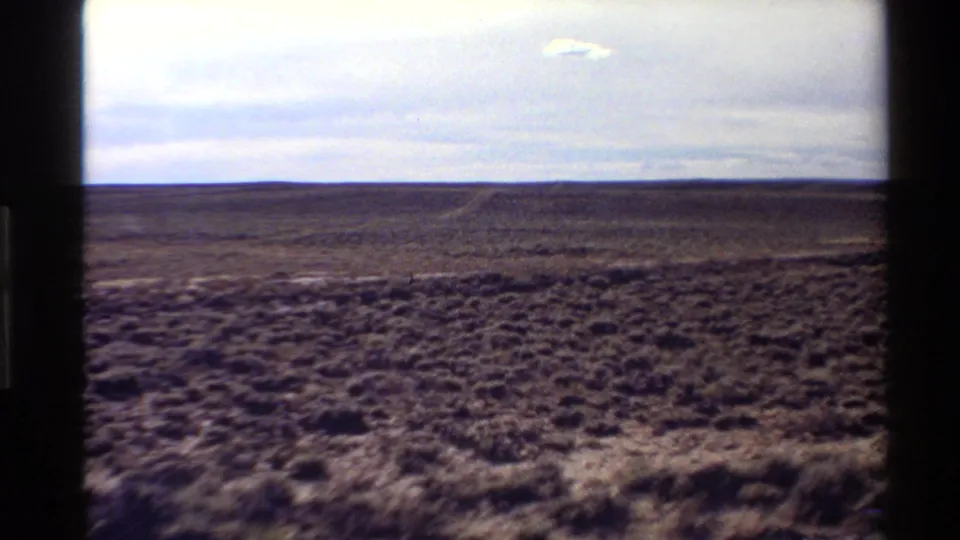 1980 Western U.S. 8mm view of open desert scrub and a low horizon beneath a cloudy sky, seen through dark frame edges.