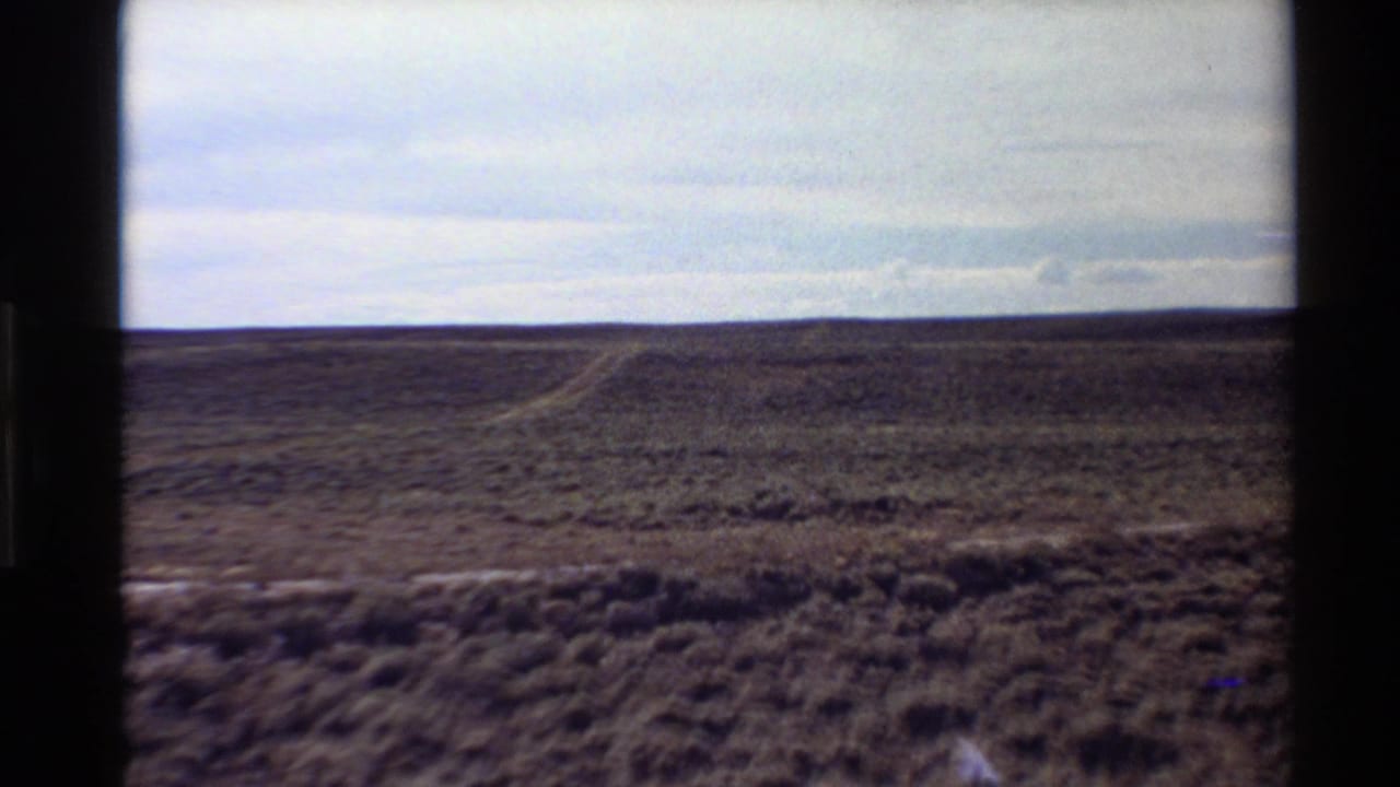 8mm home-movie frame of Western U.S. desert scrub with a pale dirt track visible below a muted cloudy sky.