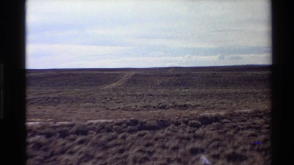 8mm home-movie frame of Western U.S. desert scrub with a pale dirt track visible below a muted cloudy sky.