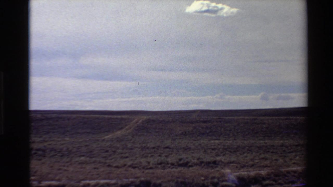 Wide 1980 Western U.S. desert view with low brush, a faint road line, cloudy sky, and dark 8mm frame borders.
