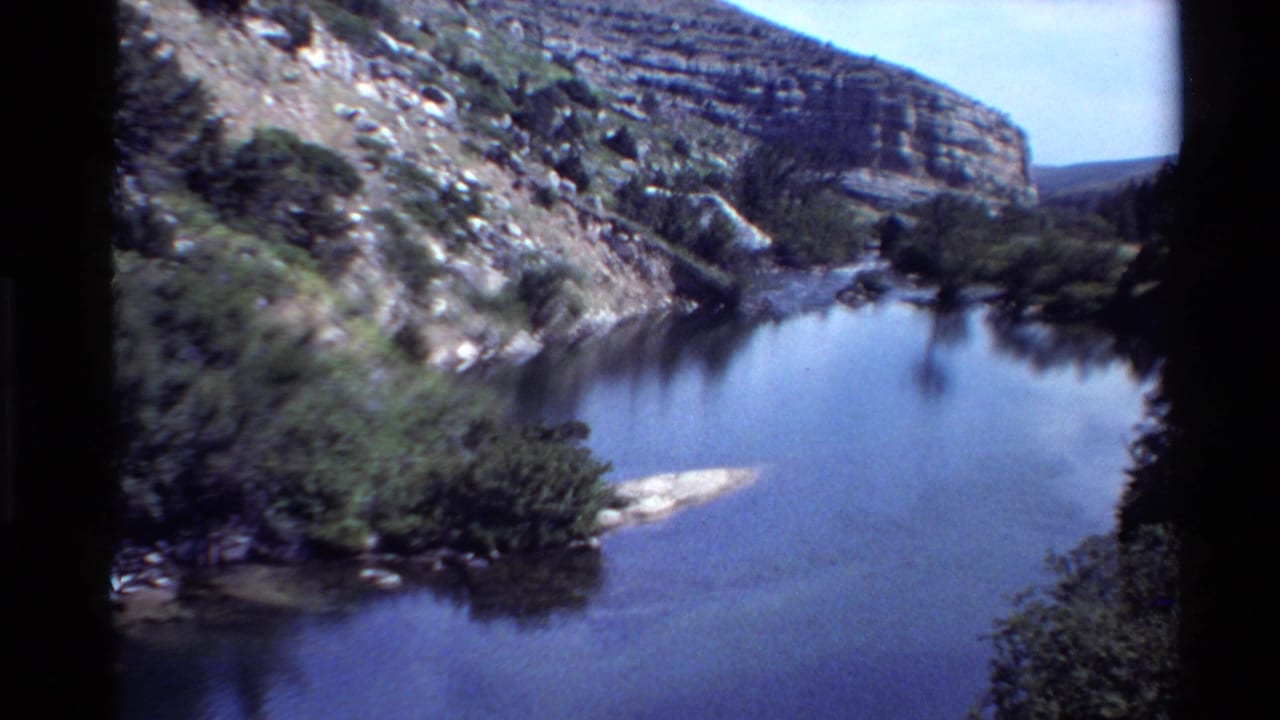 Still river water bordered by rocky canyon slopes, shrubs, and trees.