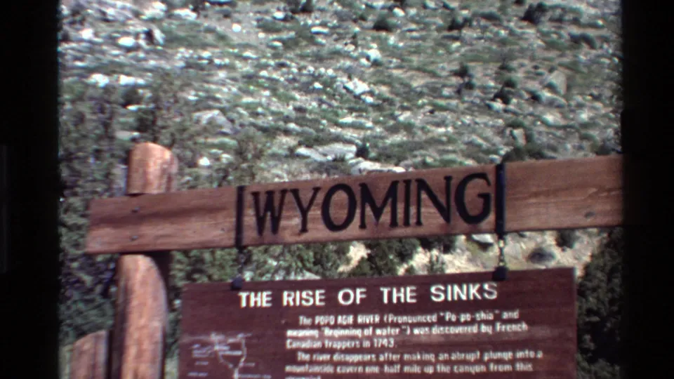 Home-movie view of the Wyoming landmark sign and lower Rise of the Sinks text panel, with rocky terrain in the background.