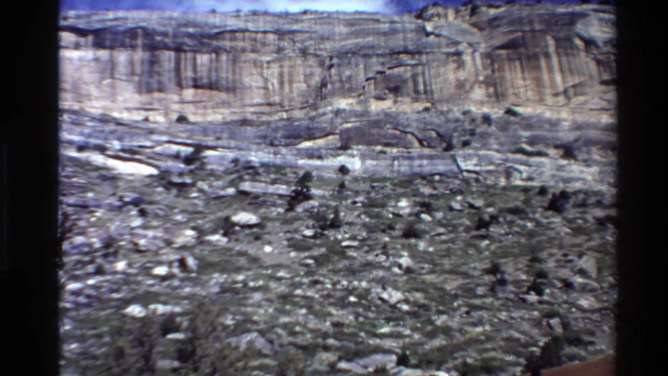 Handheld 8mm frame of scrubby rocky terrain and a banded cliff, with the sign mostly out of view.