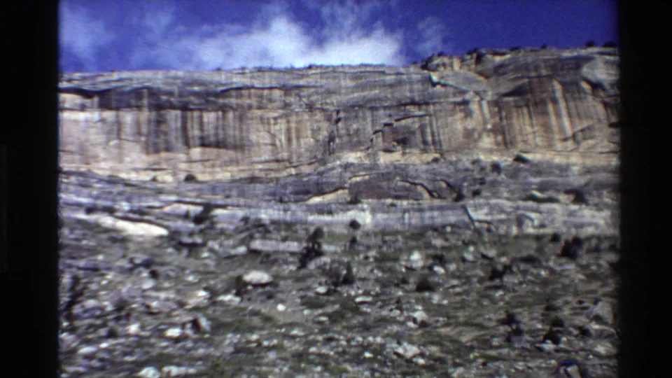 1980 Western U.S. 8mm view of a layered cliff and rocky foreground under blue sky and clouds.