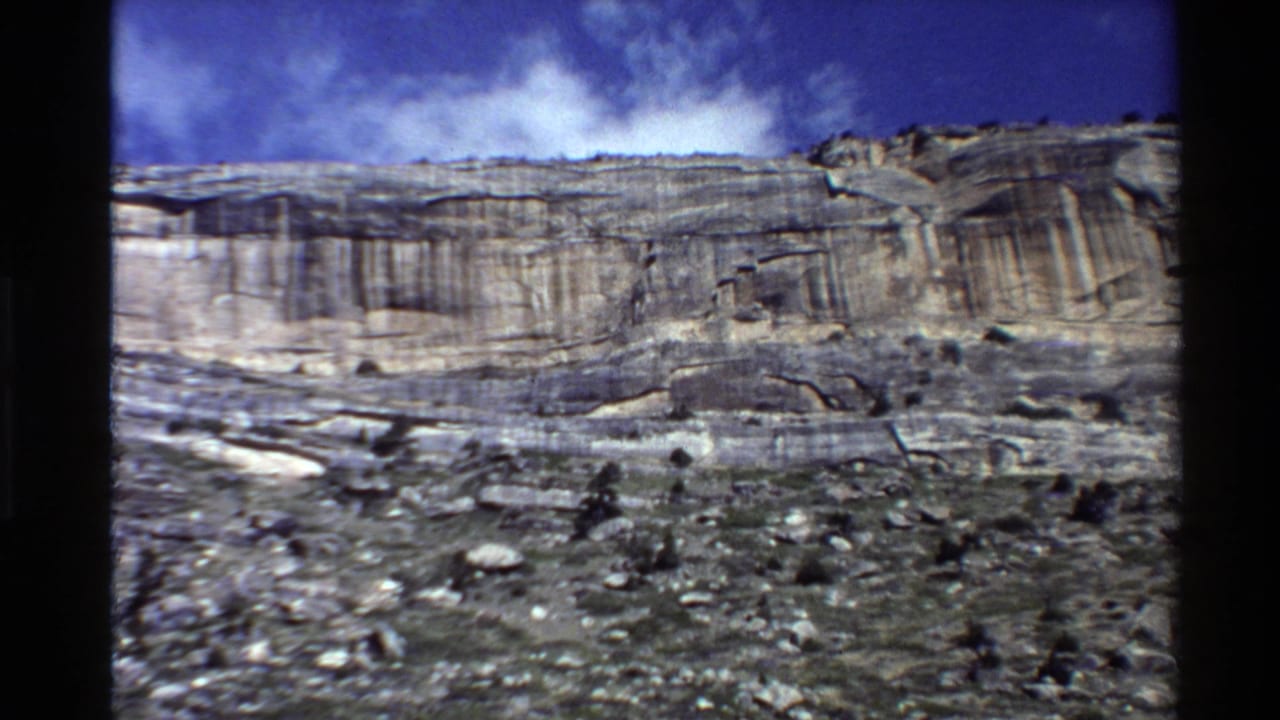 Layered rocky cliff under a blue sky with scattered clouds and rough ground in front.
