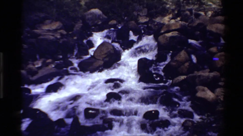 1980 Western U.S. 8mm frame of rushing white water flowing around dark rounded boulders in a rocky stream.