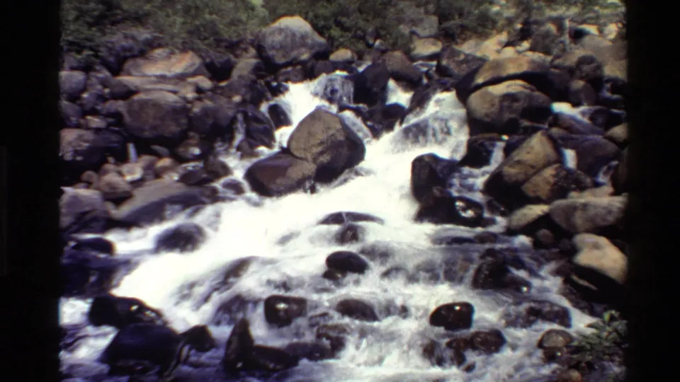 1980 Western U.S. 8mm frame showing bright white water moving through large boulders, with rocks and sparse brush in the background.