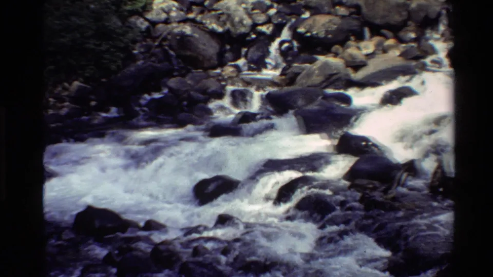 8mm footage frame of a rushing Western U.S. stream spilling across dark rocks and pale foam.