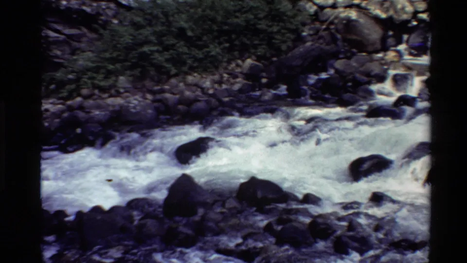 1980 home-movie frame showing a wider view of whitewater, boulders, and green foliage in the Western U.S.
