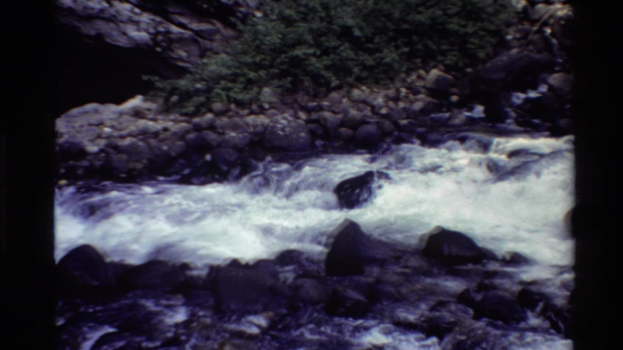 1980 8mm Western U.S. home-movie frame of a fast mountain river flowing past rocks beneath a dark stone arch.
