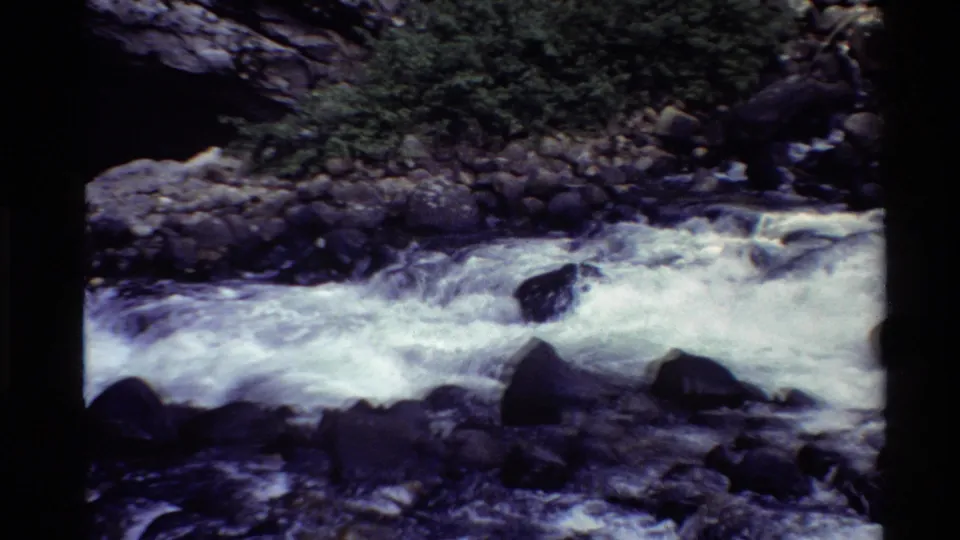 1980 8mm Western U.S. home-movie frame of a fast mountain river flowing past rocks beneath a dark stone arch.