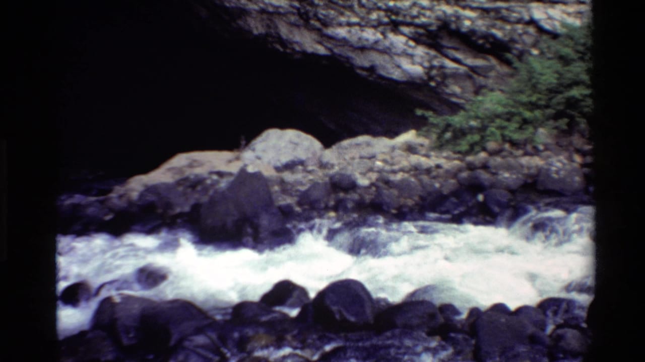 Foaming river water passes large rocks under a natural rock arch.