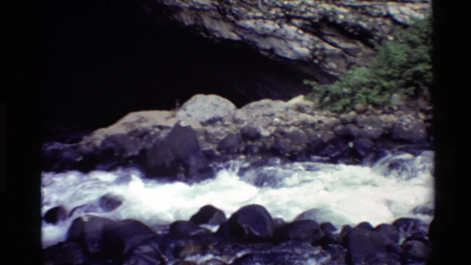 Western U.S. 8mm frame showing foaming river water and boulders below a shaded natural stone arch.