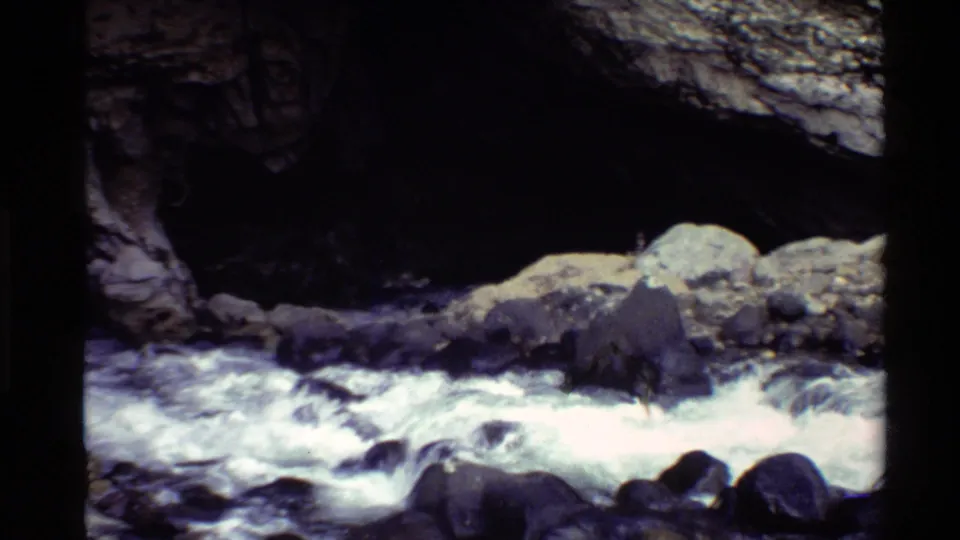 8mm home-movie view of whitewater and boulders beneath a dark stone arch in the Western United States.