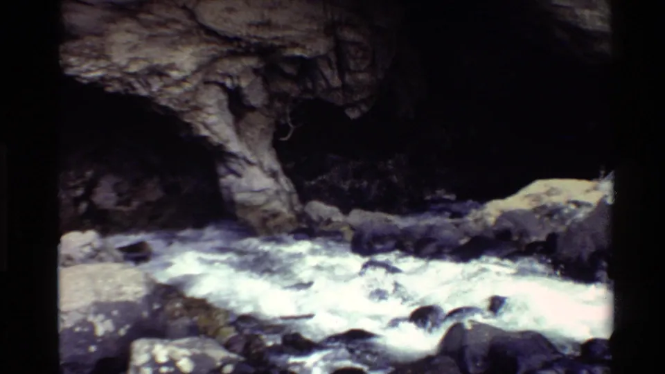 1980 archival 8mm frame of a fast river channel with whitewater, boulders, and a shaded natural stone arch.